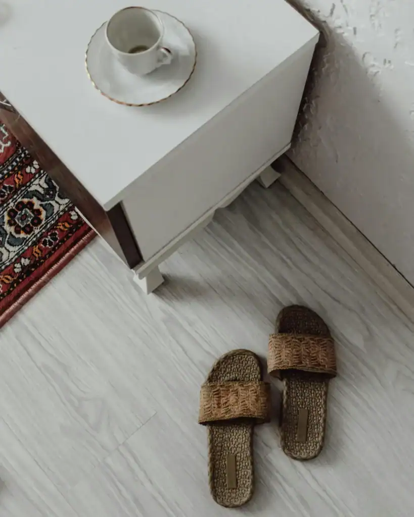 Bright whitewashed vinyl plank floor with a natural wood side table, rattan slippers, and the corner of a patterned rug