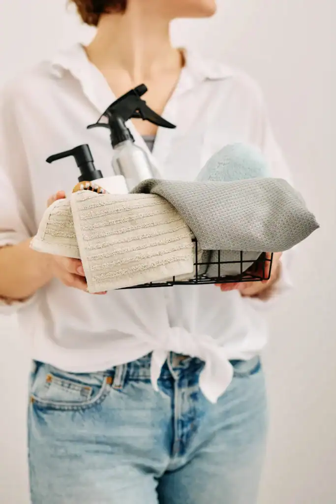 Woman holding a basket of simple supplies for a homemade diy floor cleaner, including a spray bottle, pump dispenser, and reusable cleaning cloths