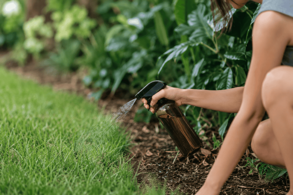 Woman watering garden with spray bottle near green plants and grass.